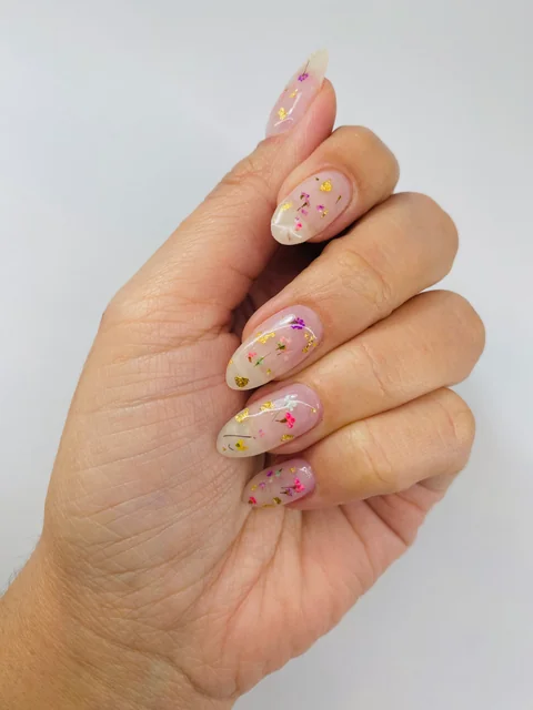 Uñas almendradas de base transparente con pequeñas flores multicolor y detalles dorados.