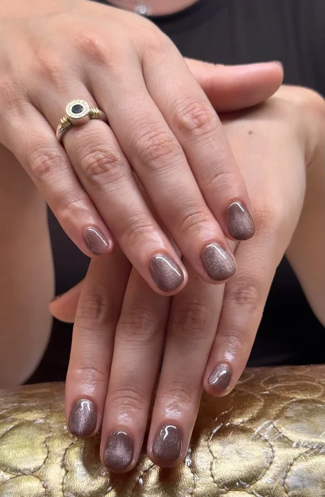 Manicura corta en tono topo con destellos sobre un apoyo dorado.